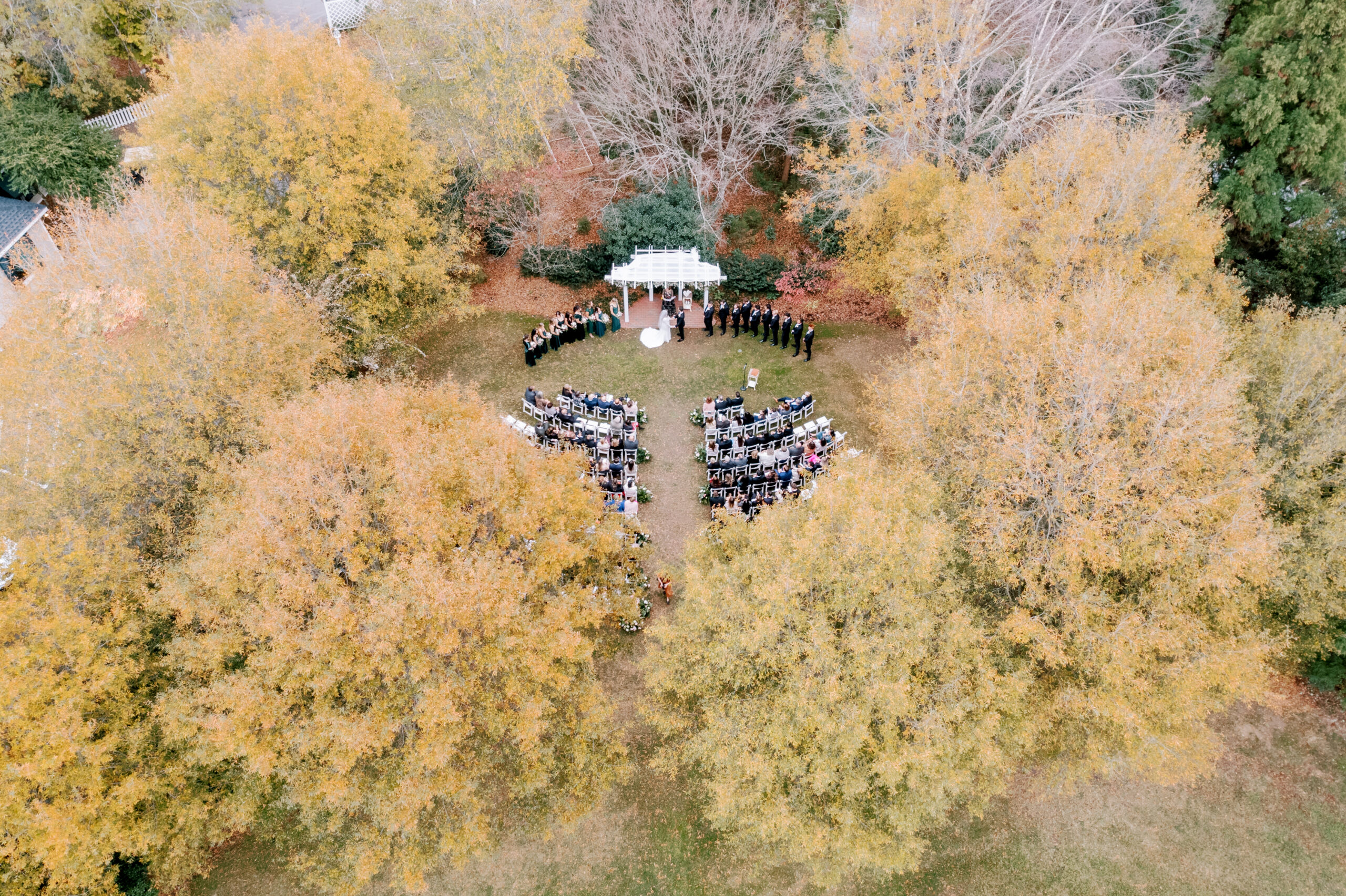 An aerial view of an outdoor wedding ceremony set among trees with golden, autumn leaves. Guests are seated in rows facing a white wedding arch where the couple stands. The scene is surrounded by vibrant fall foliage, creating a picturesque setting. Fearrington Village