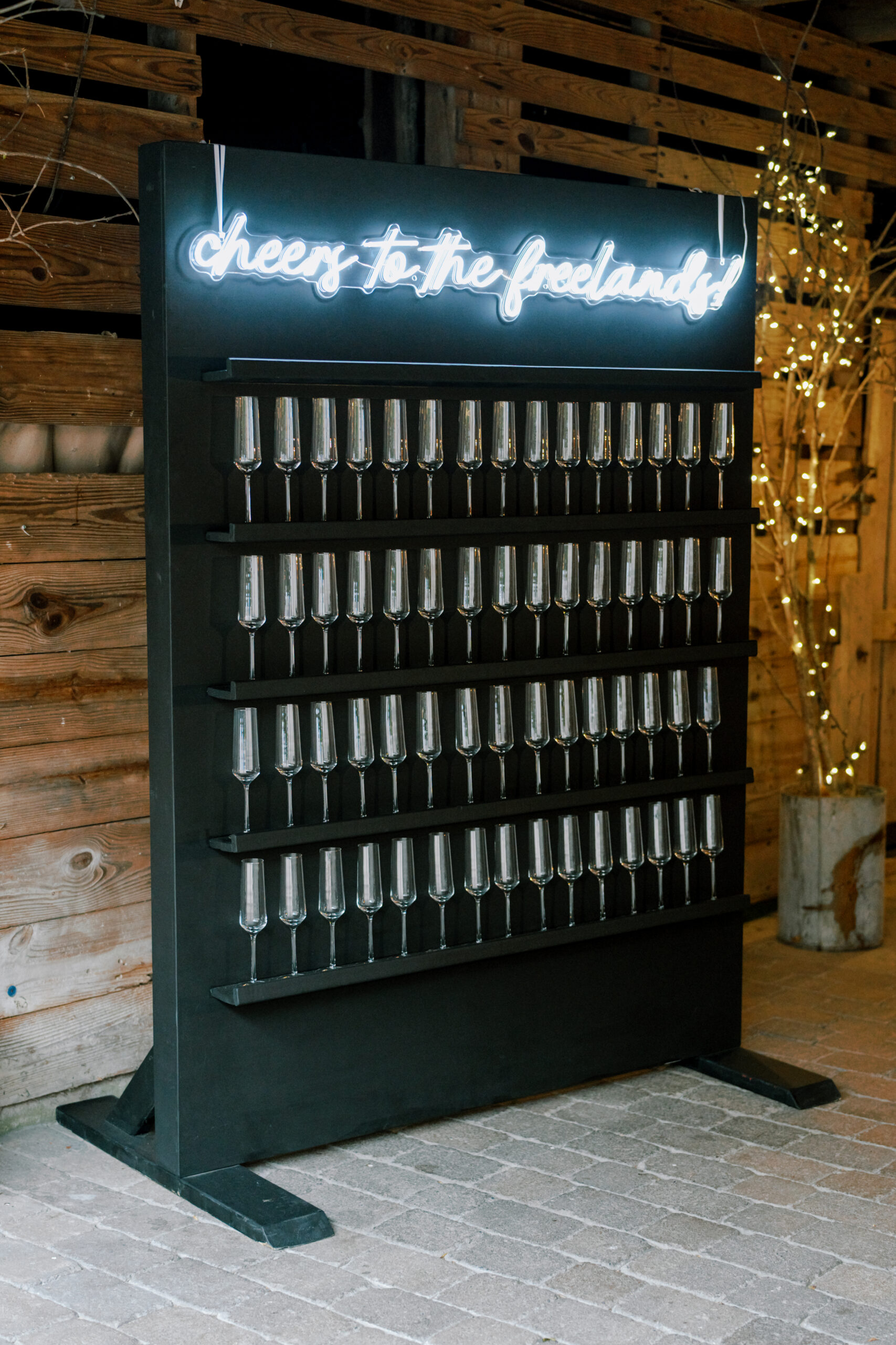 A wall display holding rows of champagne flutes, with a glowing neon sign reading "cheers to the goodlands" above it. The setup is outdoors, next to a wooden structure with decorative lights in the background. Fearrington Village