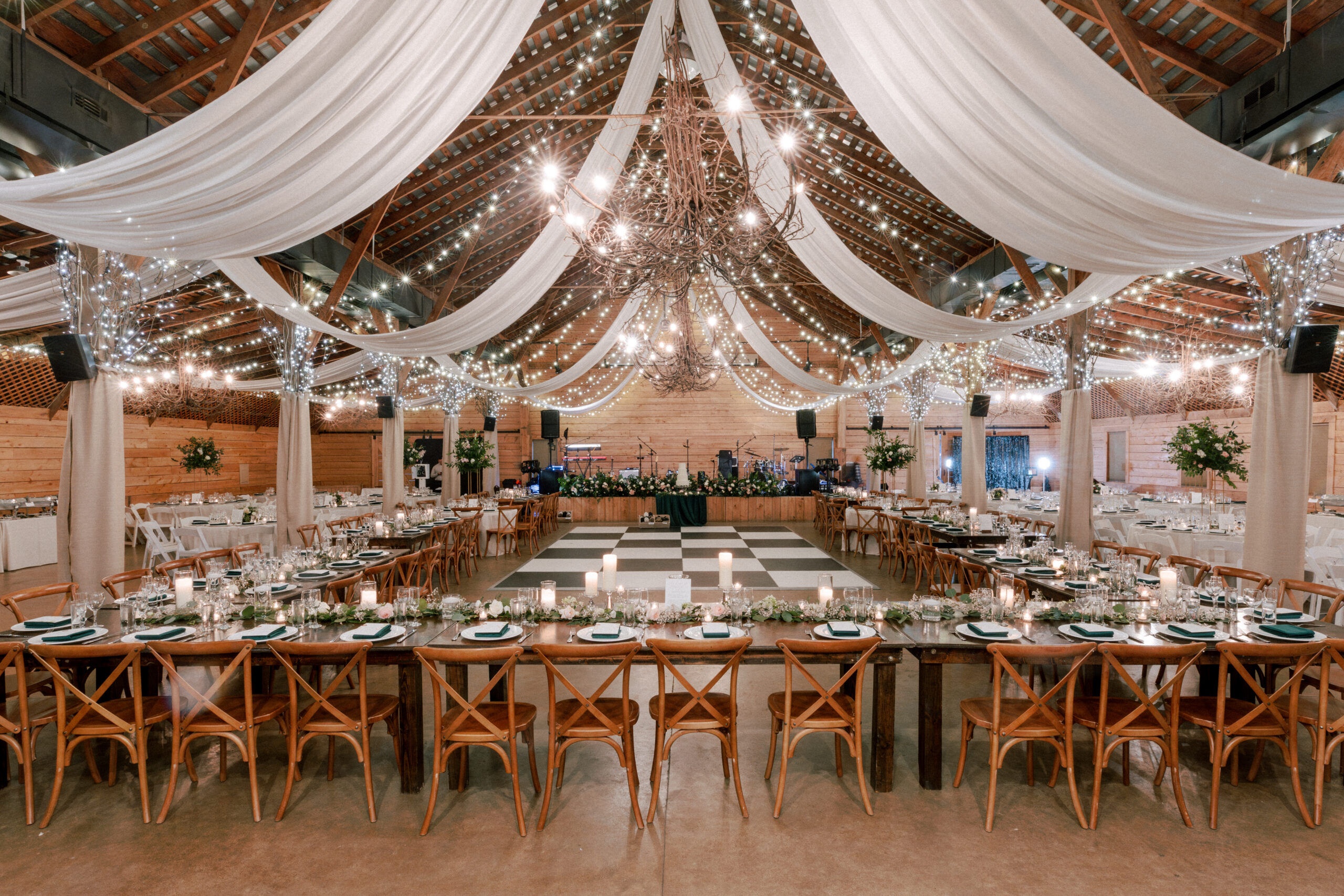 A beautifully decorated wedding venue with a rustic, wooden theme. The ceiling is adorned with draped white fabric and string lights. Long, rectangular tables are arranged in a U-shape around a central dance floor, set with elegant tableware and candles. Fearrington Village