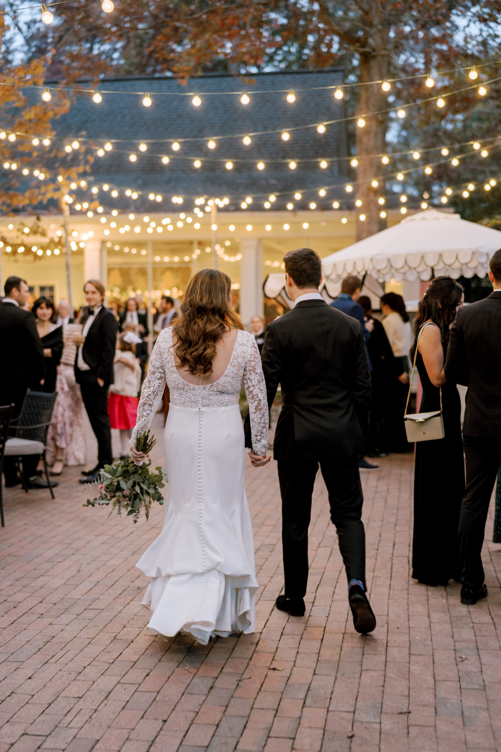 A bride and groom walk hand-in-hand on a brick path at a wedding reception under string lights. The bride, in a white gown with lace sleeves, holds a bouquet, while the groom wears a black suit. Guests in formal attire socialize in the background. Fearrington Village