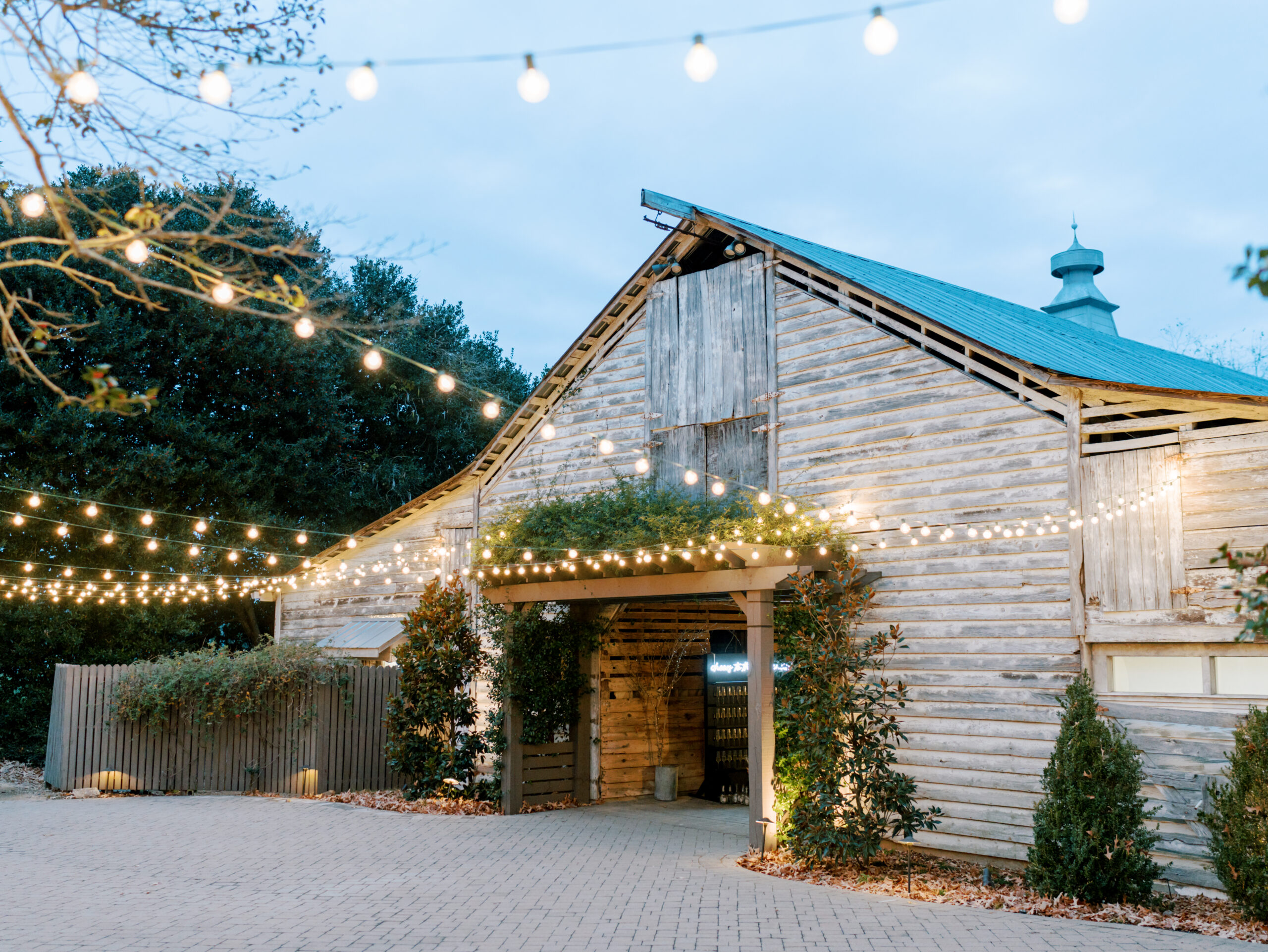 The Fearrington Barn. A rustic wooden barn is adorned with string lights, creating a warm and inviting atmosphere. The barn has a metal roof and is surrounded by trees and bushes. The ground is paved, and the lights cast a soft glow, suggesting an evening or nighttime setting. Fearrington Village