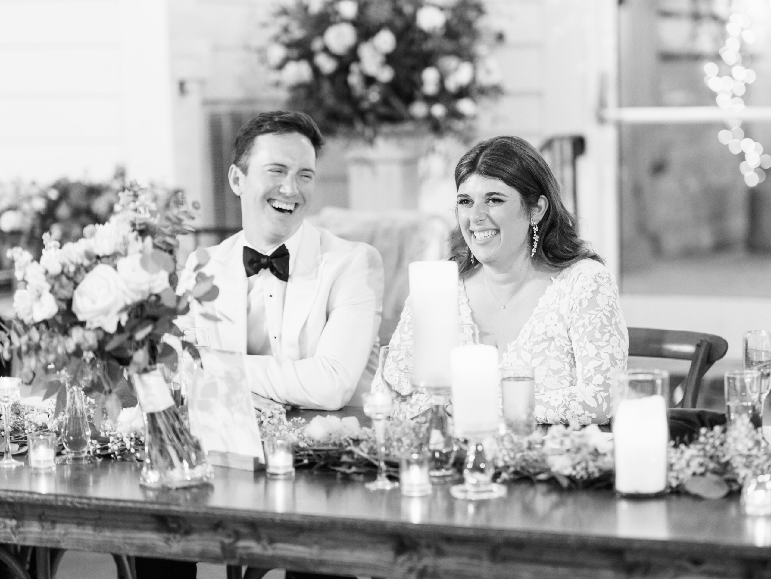 A bride and groom sit at a wooden table adorned with floral arrangements and candles. Both are laughing heartily. The groom wears a white tuxedo jacket with a black bow tie, and the bride is in a lace wedding dress. Blurred background suggests a festive setting. Fearrington Village