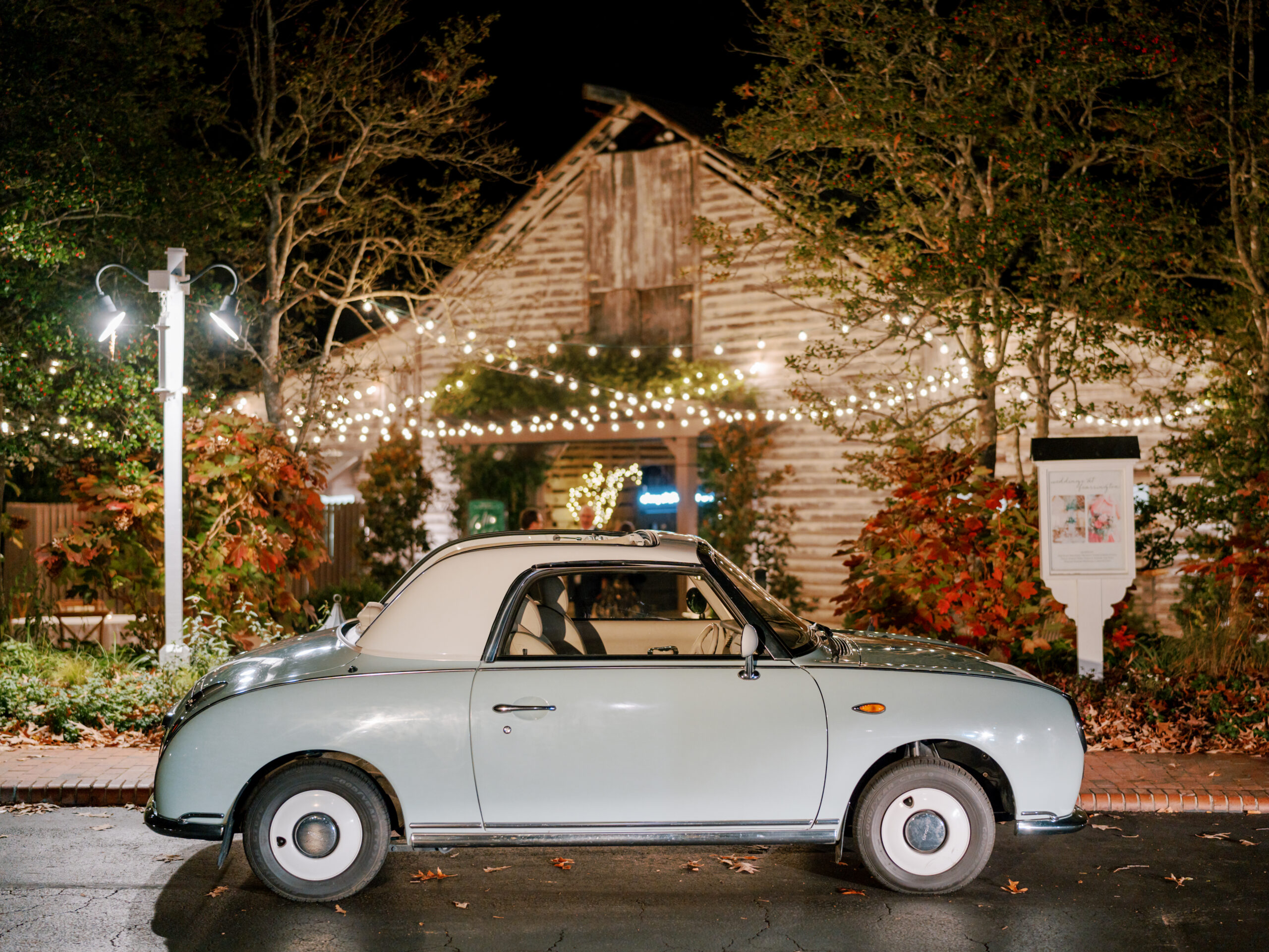 A vintage light blue car with a white roof is parked on a wet street at night, in front of a rustic building adorned with string lights and surrounded by trees with autumn leaves. A well-lit signboard is visible to the right of the car. Fearrington Village