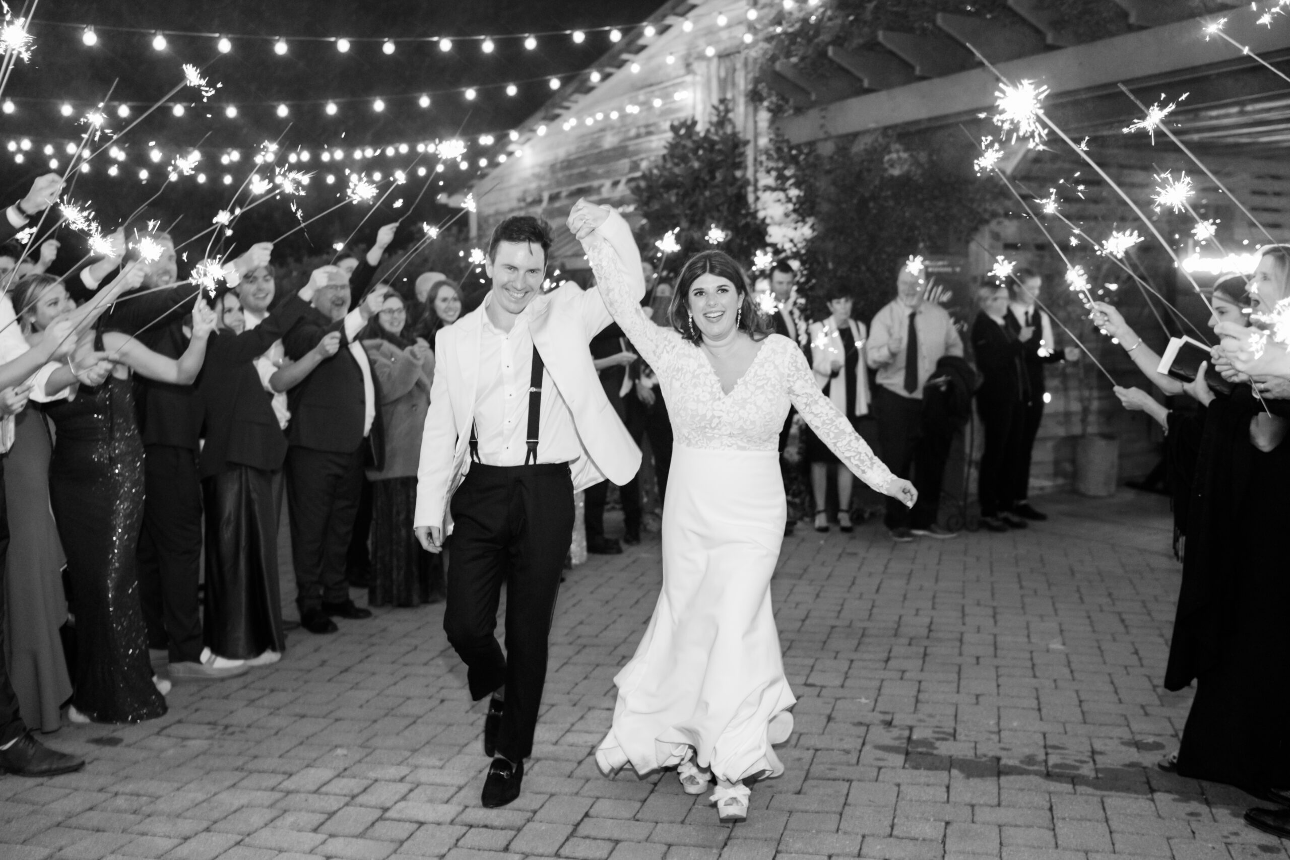 A bride and groom, both smiling, walk hand in hand under a canopy of string lights. The groom wears a white jacket and suspenders while the bride dons a long-sleeve lace gown. Guests surround them, holding lit sparklers and celebrating their exit. Fearrington Village