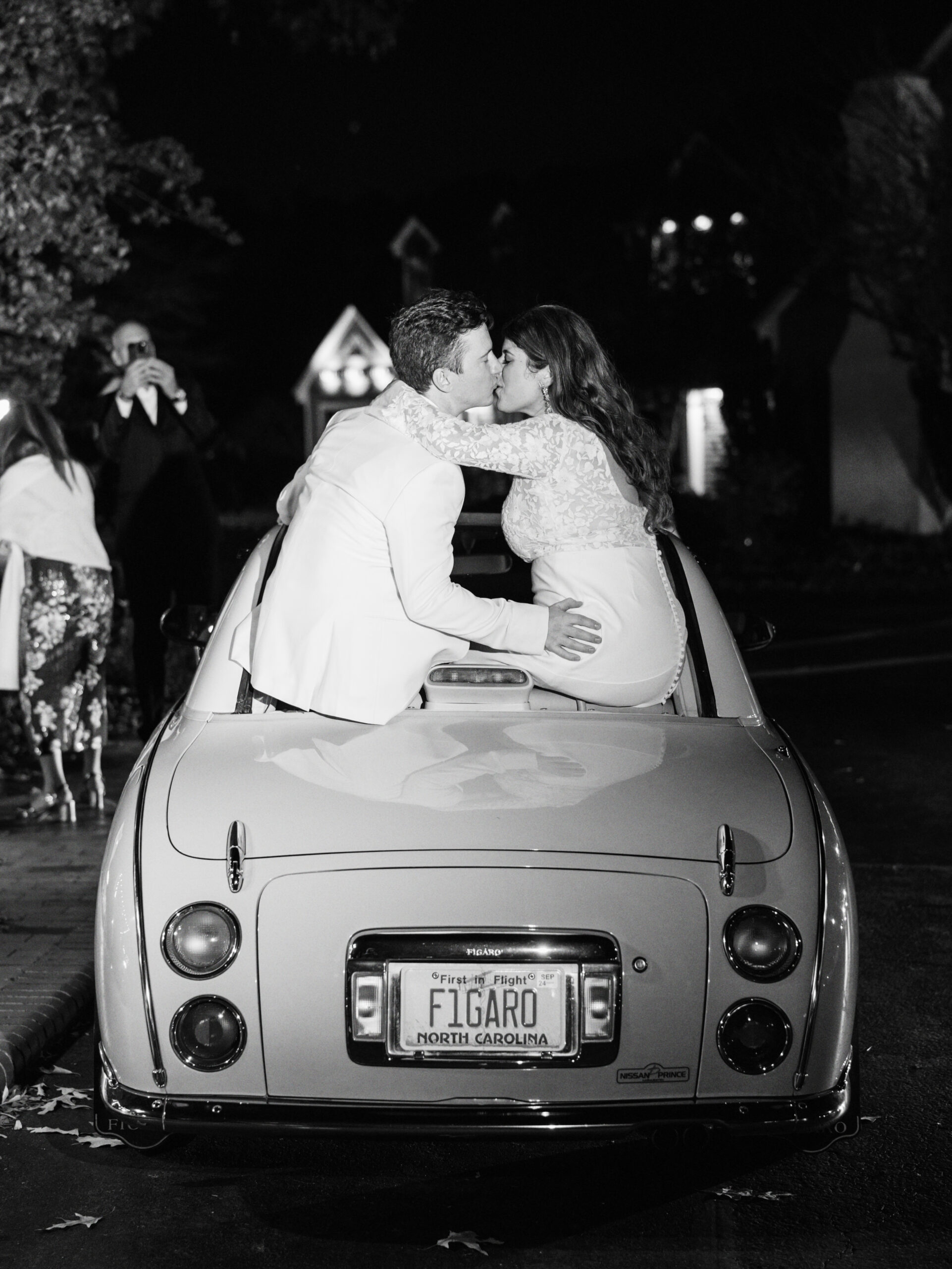 A couple, dressed in wedding attire, share a kiss while standing on the back of a vintage convertible car at night. The car's license plate reads "FIGARO" and indicates North Carolina. Onlookers nearby capture the moment with their phones. Fearrington Village