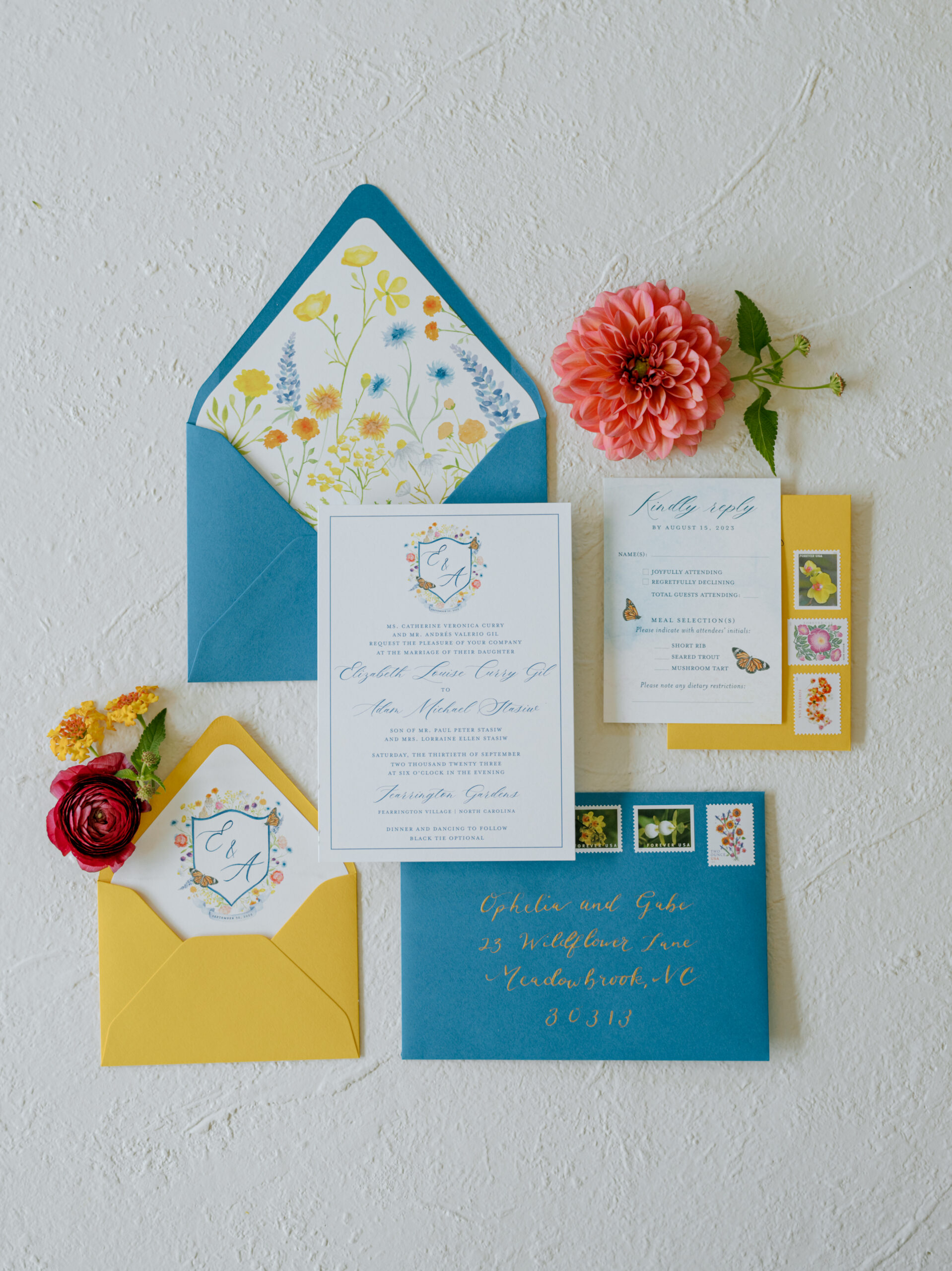 A flat lay image of a wedding invitation set on a white surface. The set includes a blue envelope, yellow envelope, RSVP card, invitation card, and floral stamps. A red rose, yellow carnation, and pink dahlia decoratively surround the stationary. Fearrington Village