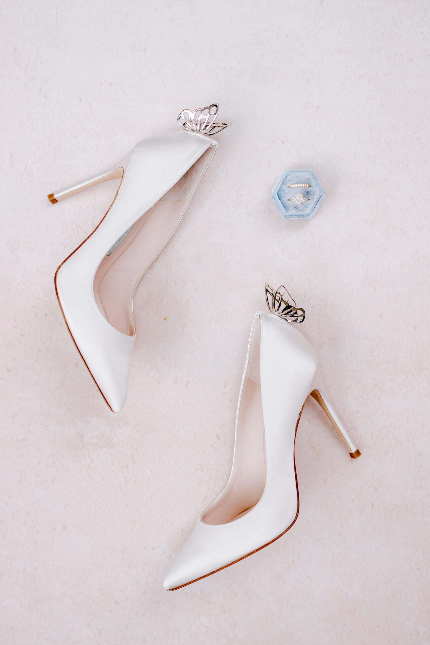 A pair of white high-heeled shoes featuring silver butterfly ornaments on a light surface. Near the shoes, there is a hexagonal blue ring box with a ring inside. Fearrington Village