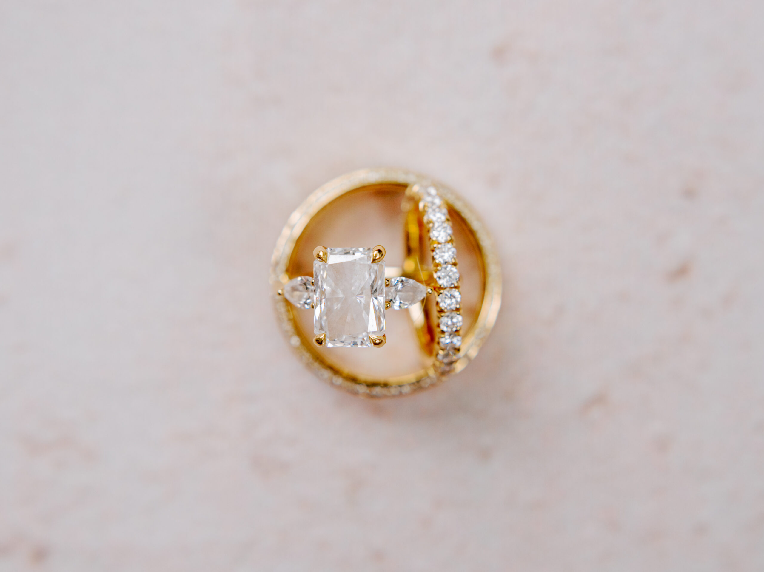 A close-up image of two rings on a light, textured background. One ring features a large rectangular diamond flanked by two smaller stones on a gold band. The other is a gold band encrusted with small round diamonds. The rings are arranged in a circular formation. Fearrington Village