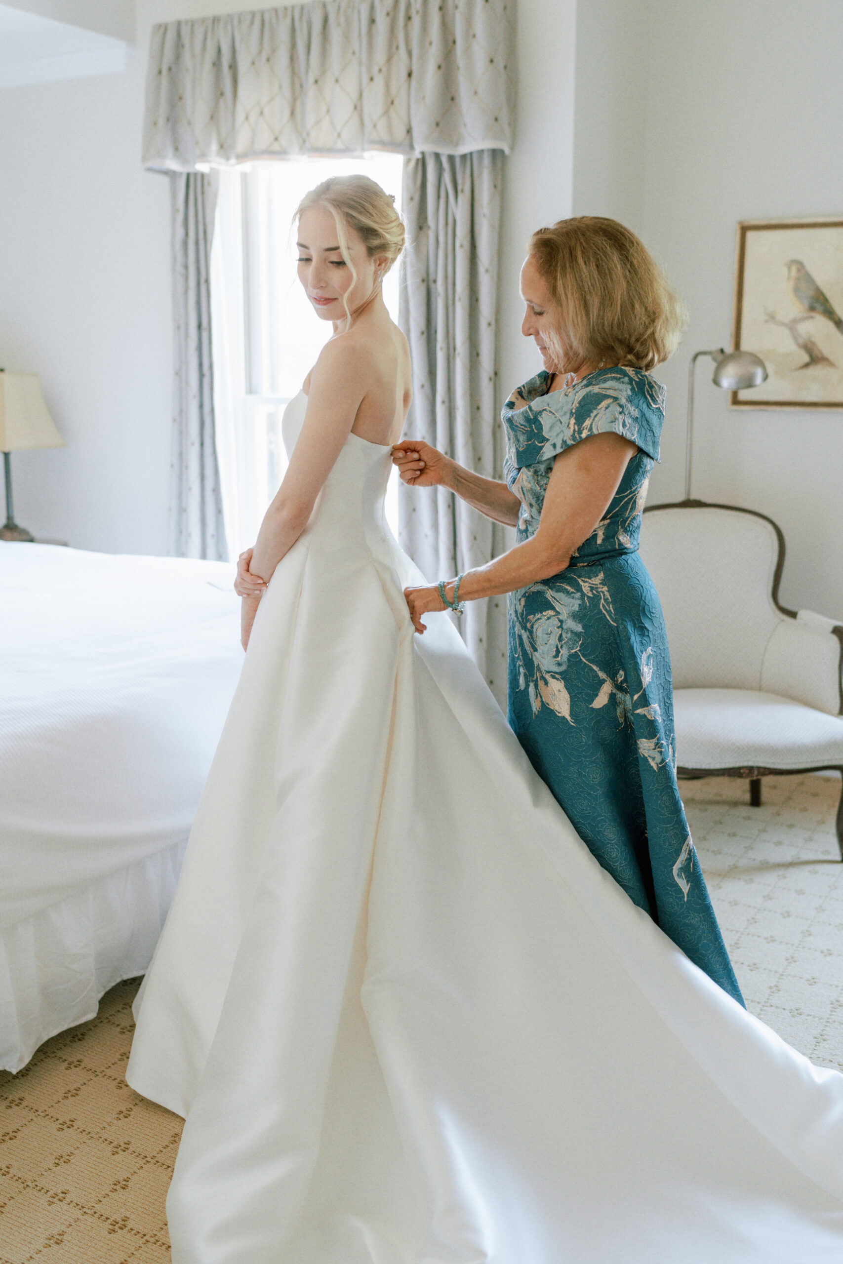 A bride in a strapless white gown stands by a bed as a woman in a blue dress helps adjust the back of her dress. The room features white walls, a framed bird artwork on the wall, and a curtain-covered window letting in natural light. Fearrington Village