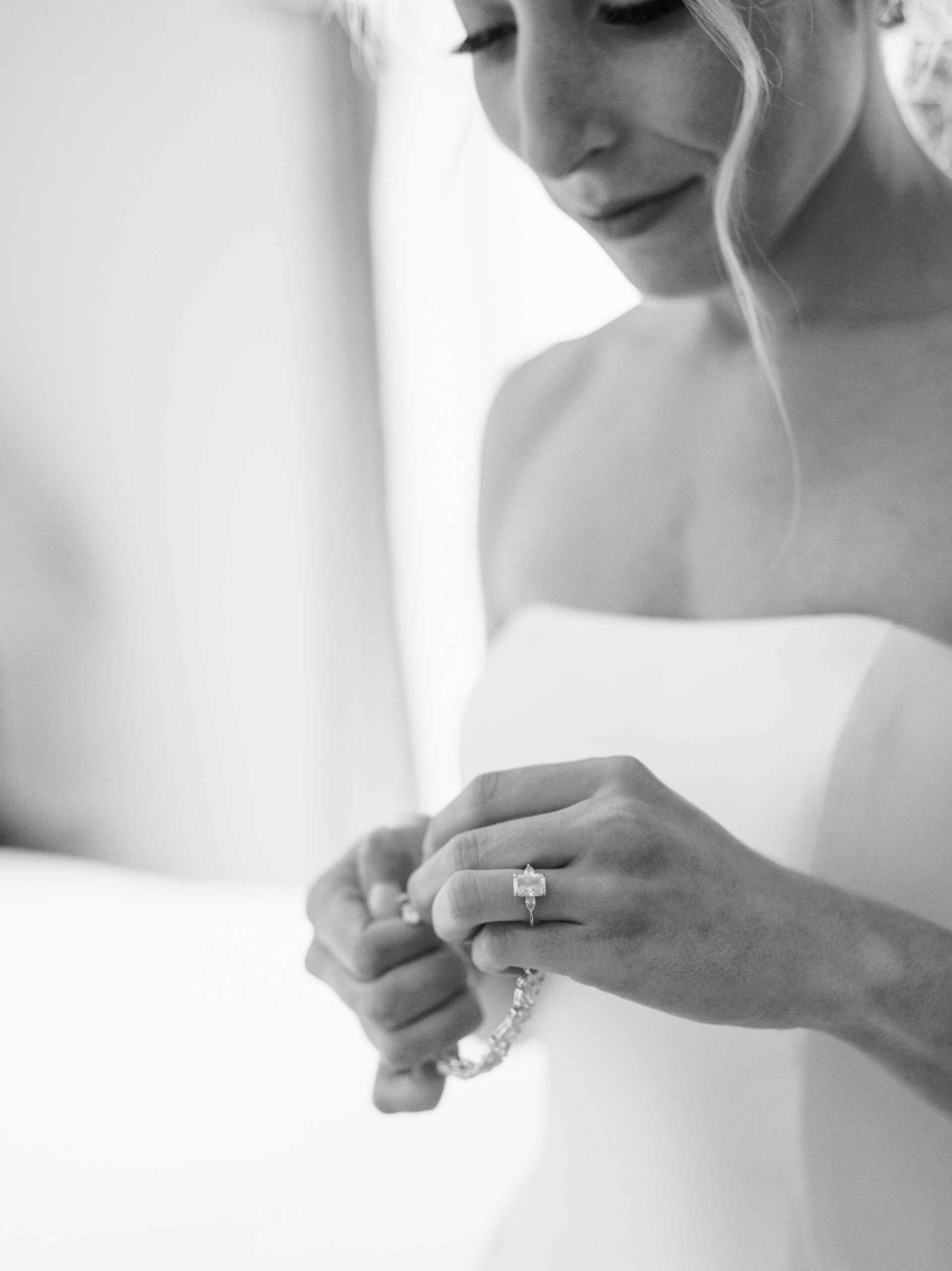 Black and white photo of a bride in a strapless wedding dress, looking down, and holding her bracelet. Her engagement ring is visible on her left hand. The background is softly blurred, focusing attention on the bride's serene and contemplative expression. Fearrington Village