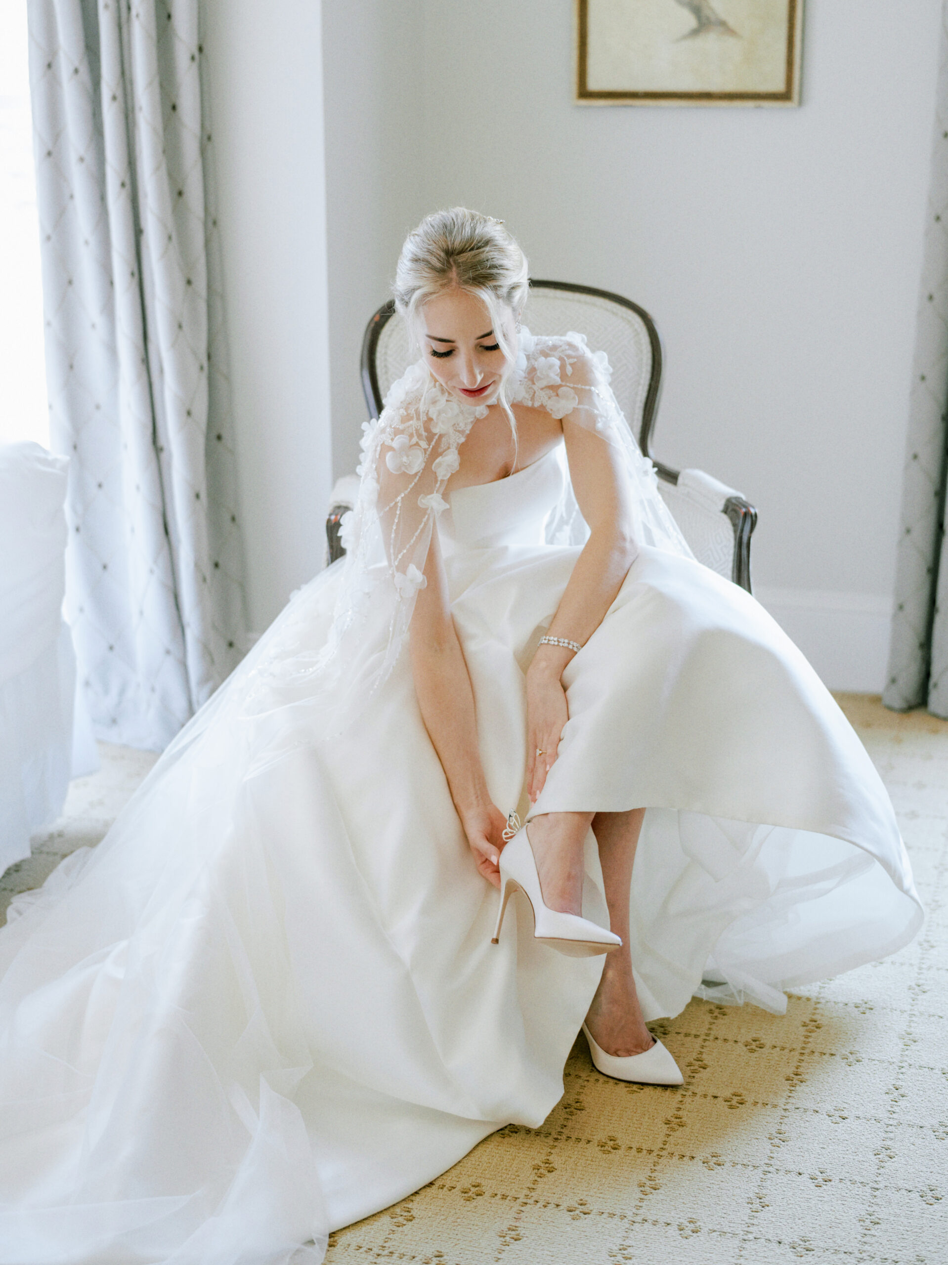 A bride sits on a chair in a well-lit room while adjusting the strap of her white high-heeled shoe. She is wearing a white wedding gown with intricate floral detailing on the shoulders. The room features light-colored walls, curtains, and a framed bird artwork. Fearrington Village