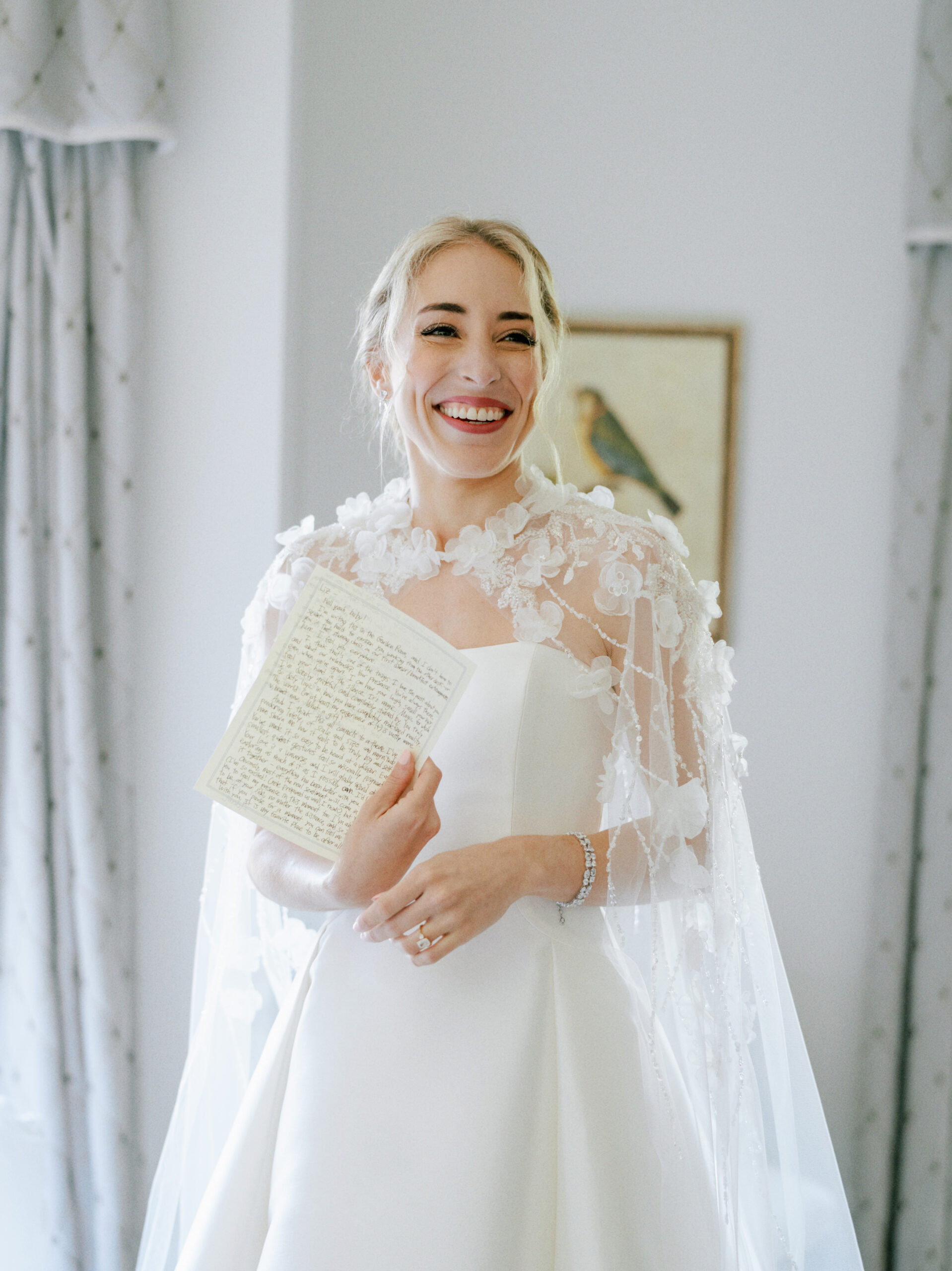 A smiling bride holding a handwritten letter stands in a softly lit room. She is wearing a white wedding dress with an intricate lace overlay. Light-colored curtains frame the window behind her, and a bird illustration is visible on the wall in the background. Fearrington Village