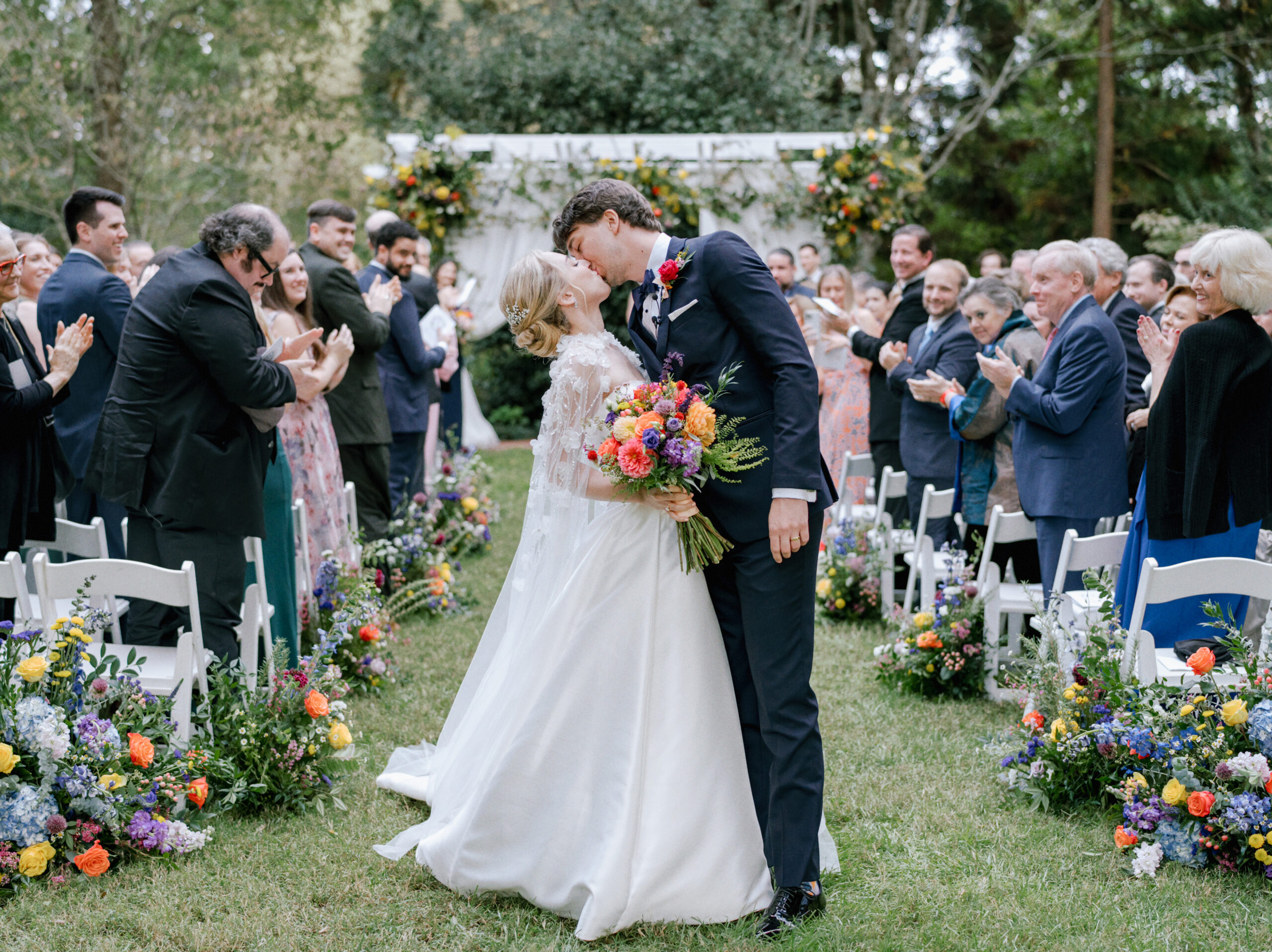 A newlywed couple shares a kiss in the middle of an outdoor aisle adorned with colorful flowers. The bride holds a vibrant bouquet while guests on both sides, dressed in formal attire, clap and celebrate. A floral archway decorates the background. Fearrington Village