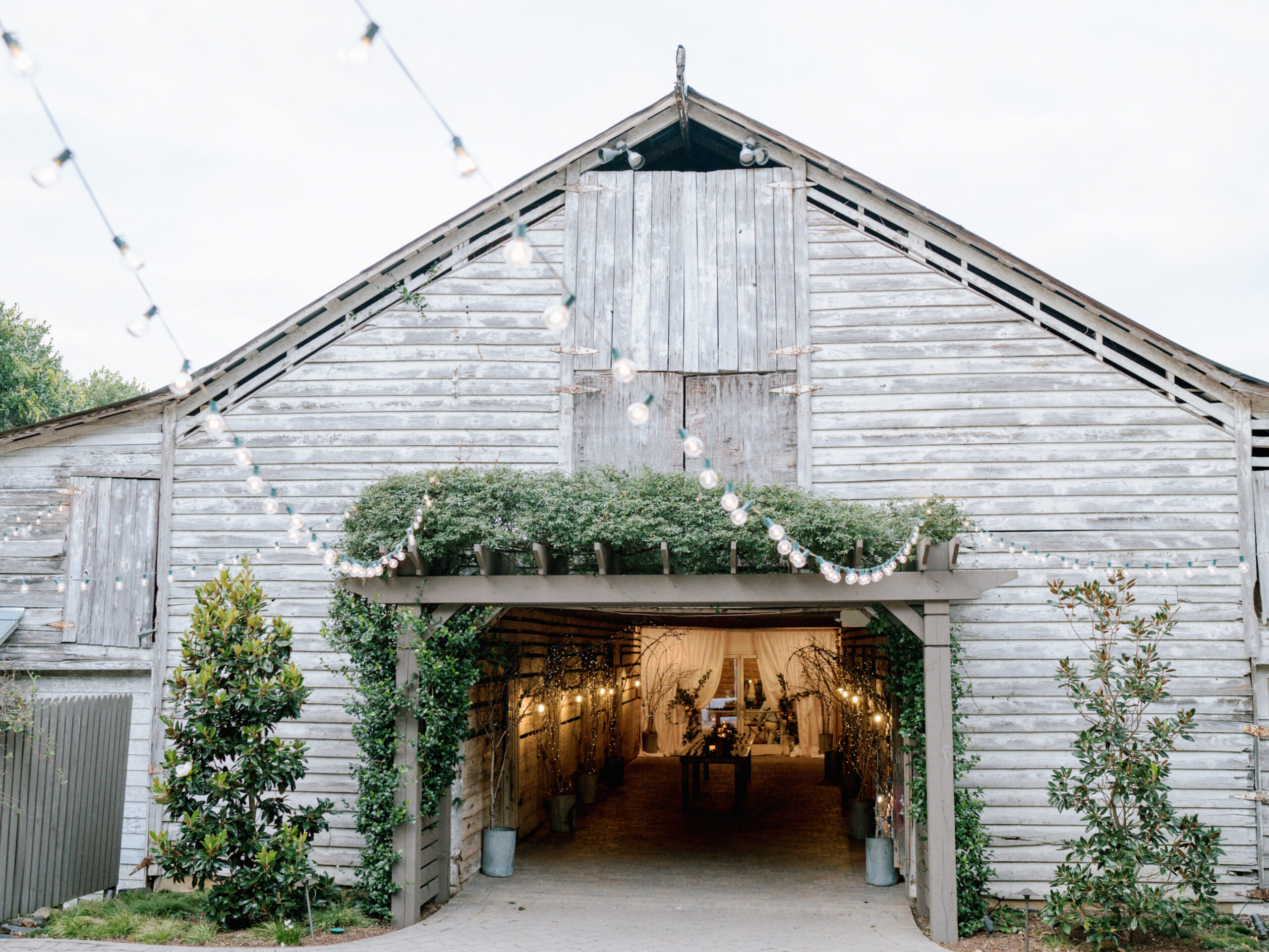 A rustic, weathered barn adorned with string lights and green foliage, creating a cozy and inviting entrance. The interior is softly illuminated, suggesting an event or gathering inside. Potted plants flank the entrance, adding a touch of greenery. Fearrington Village