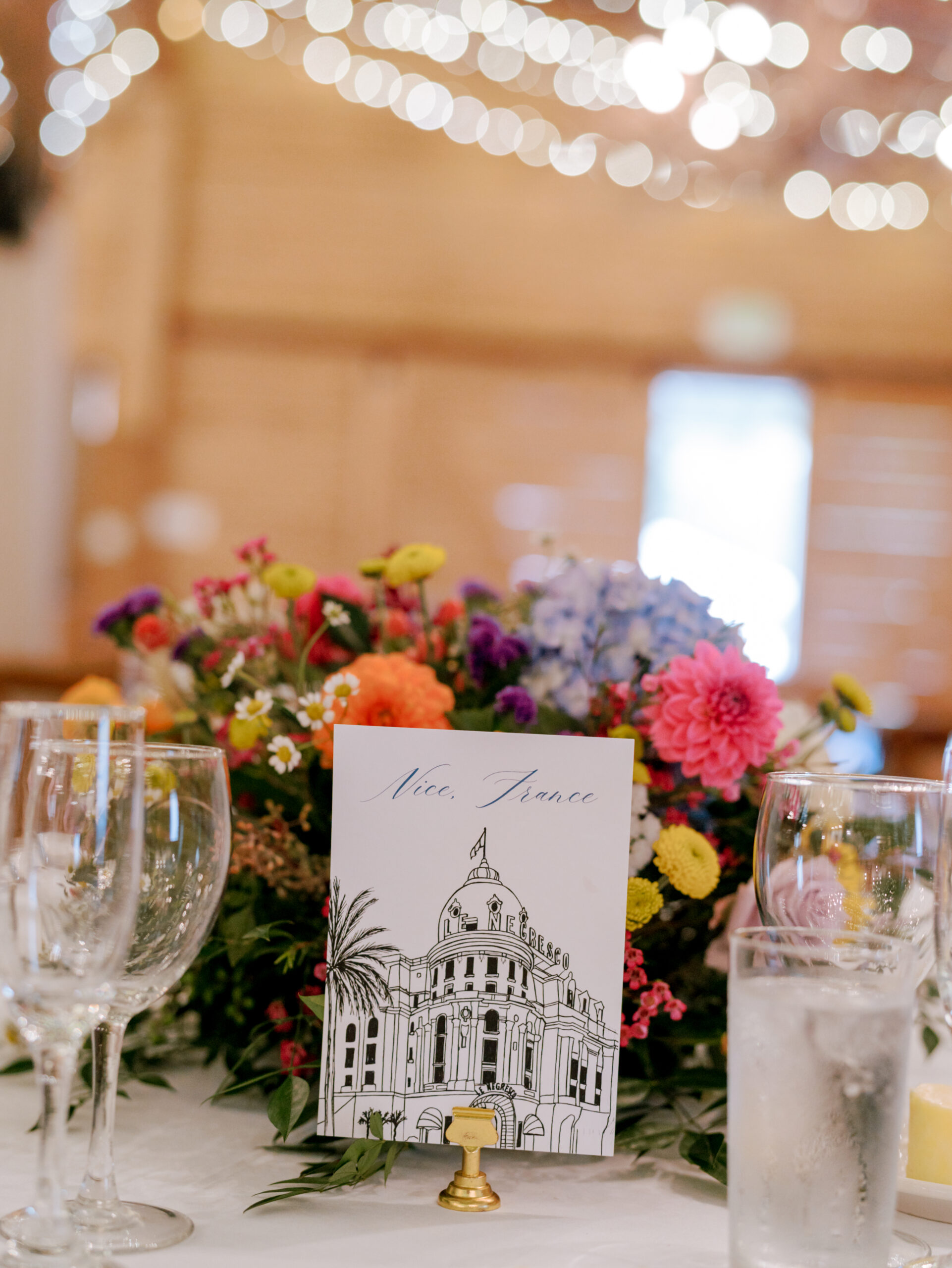 A table decoration featuring a bouquet of colorful flowers and a card with an illustration of a building in Nice, France. Surrounding the bouquet are wine glasses and a glass of water, with twinkling lights softly visible in the background. Fearrington Village