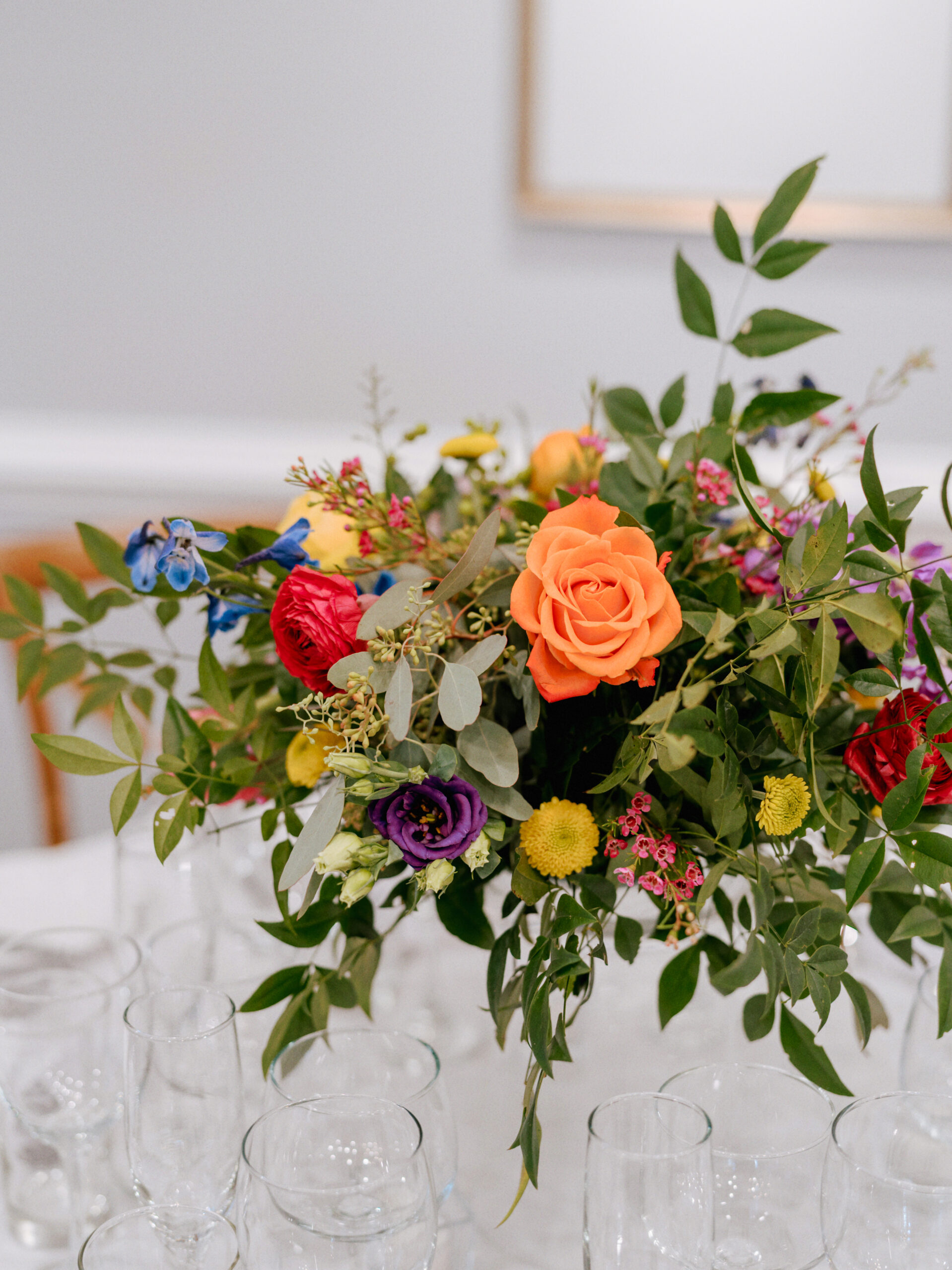 A vibrant floral arrangement features a variety of colorful flowers, including an orange rose as the centerpiece, surrounded by greenery, purple lisianthus, yellow button poms, and other blooms. Clear glassware is arranged on the table beneath the bouquet. Fearrington Village