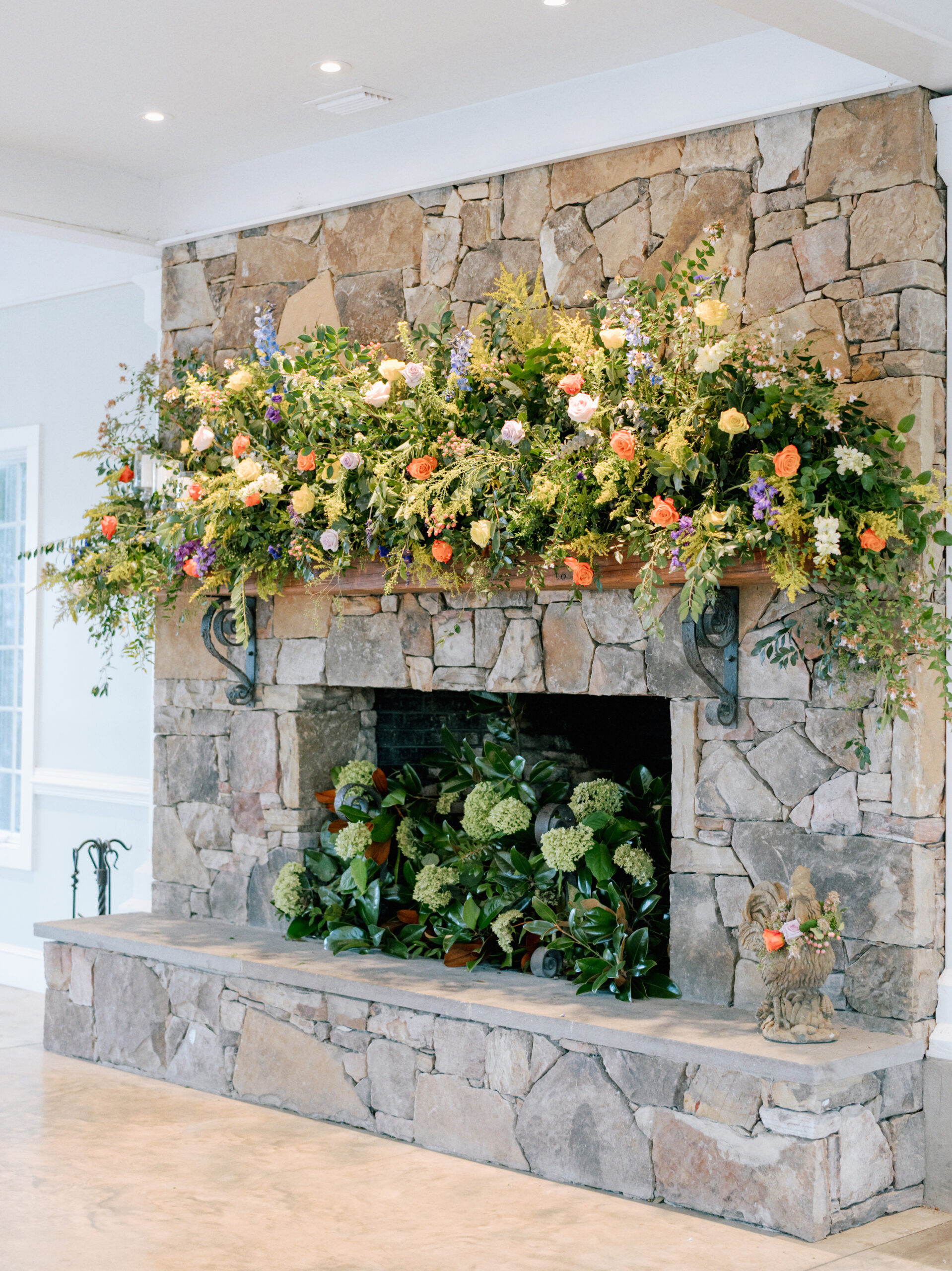 A rustic stone fireplace adorned with abundant greenery and colorful flowers. The floral arrangement extends across the mantle, featuring shades of orange, yellow, lilac, and white. Additional green foliage decorates the hearth, creating a lush, vibrant display. Fearrington Village