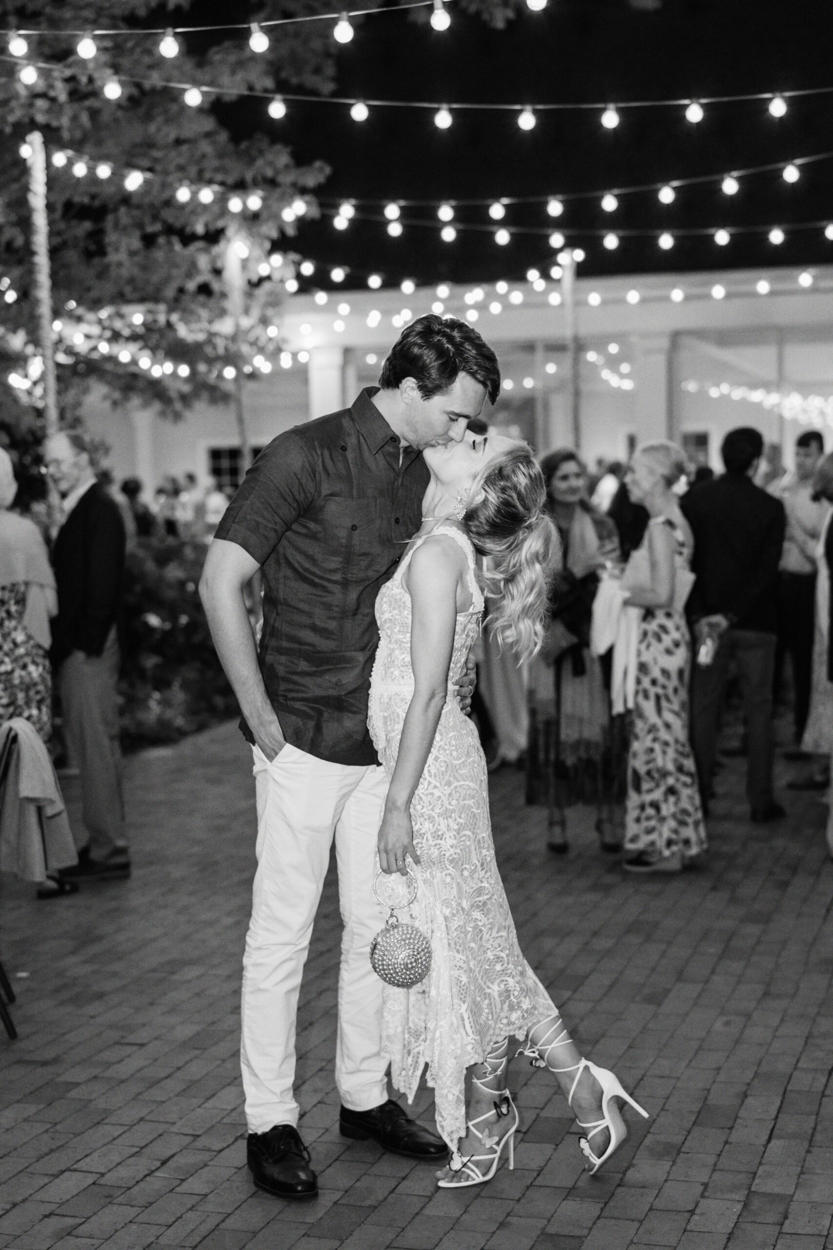 A couple shares a kiss at an outdoor evening event, illuminated by string lights. The man, wearing a dark shirt and light pants, has his hand in his pocket. The woman, in a lace dress and heels, holds a small round purse. People mingling can be seen in the softly lit background. Fearrington Village