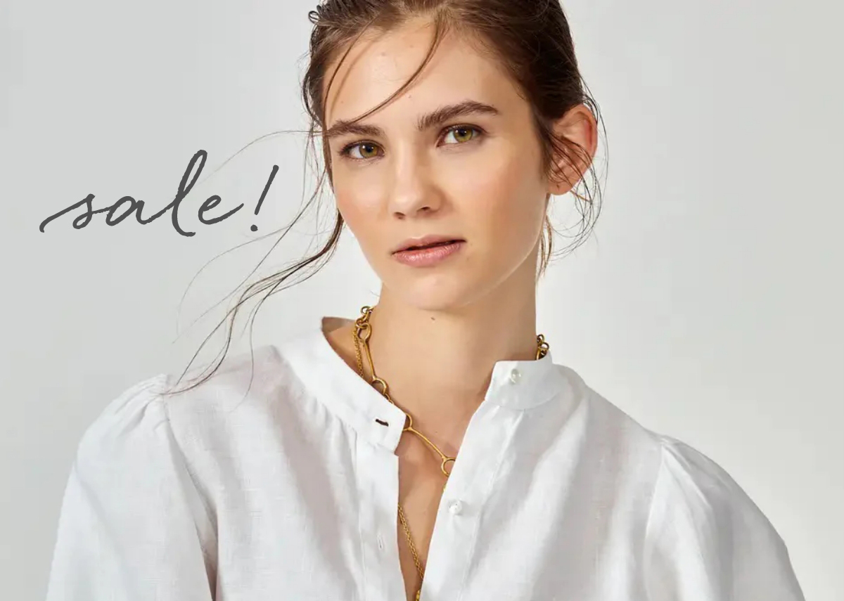A woman with tied-back brown hair and loose strands framing her face is wearing a white blouse and a gold chain necklace. She is looking directly at the camera with a neutral expression. The word "sale!" is written in a stylish font to the left of her. Fearrington Village