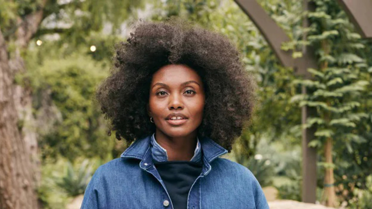 A person with a voluminous afro hairstyle stands outdoors in a natural setting. They are wearing a blue denim jacket over a dark top. The background features lush greenery and an arch-shaped wooden structure. Fearrington Village