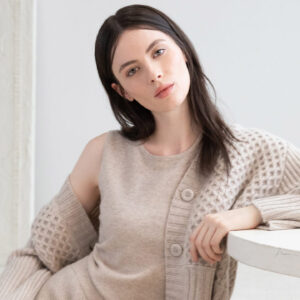 A woman with long dark hair is seated beside a round white table. She is wearing a beige knit sweater with a pattern and a matching sleeveless top. Her gaze is directed at the camera and she has a relaxed posture. The background is a light-colored, textured wall. Fearrington Village