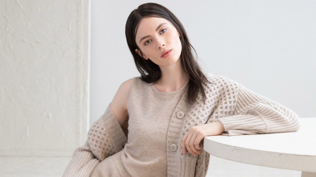 A woman with long dark hair is seated beside a round white table. She is wearing a beige knit sweater with a pattern and a matching sleeveless top. Her gaze is directed at the camera and she has a relaxed posture. The background is a light-colored, textured wall. Fearrington Village