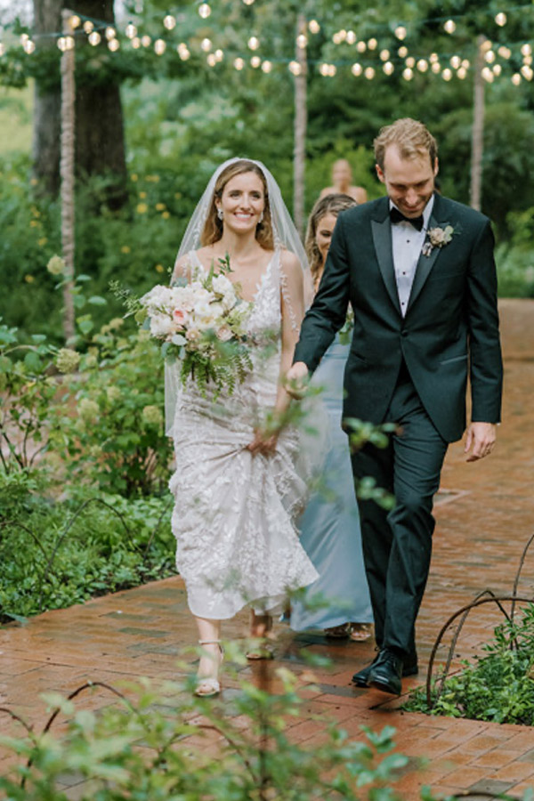 A bride and groom walk down an outdoor path lined with greenery. The bride, smiling and holding a bouquet of white and pink flowers, wears a white lace gown and veil. The groom, in a dark suit with a bow tie, looks down while holding her hand. Twinkling string lights hang above. Fearrington Village