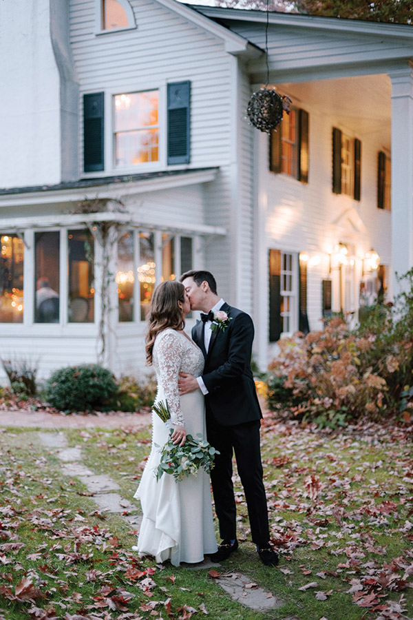 A bride and groom share a kiss in front of a picturesque white house with black shutters. The bride is in a long, white gown holding a bouquet, while the groom wears a black suit and bow tie. Autumn leaves are scattered on the ground around them. Fearrington Village