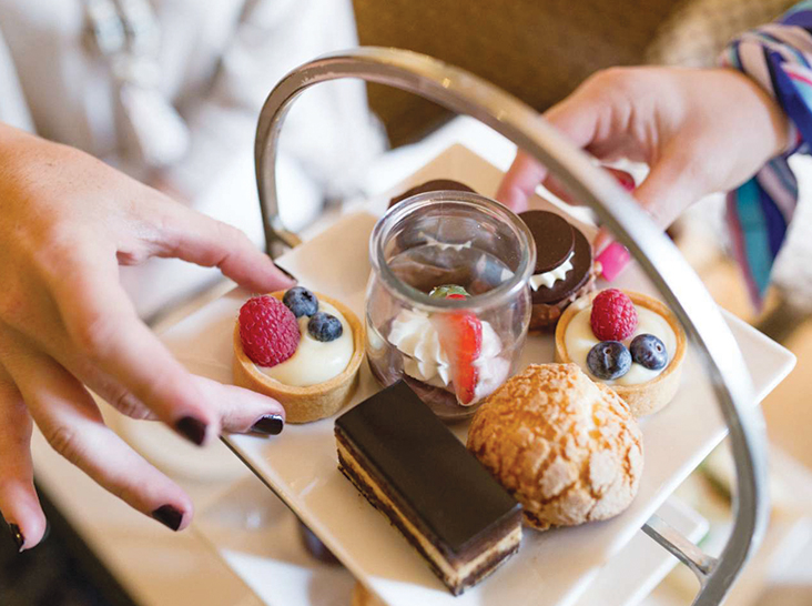 A top view of a dessert tray with various small pastries, including tarts topped with raspberries and blueberries, a chocolate eclair, and a glass dessert with whipped cream and strawberries. Two hands are picking sweets from the tray. Fearrington Village