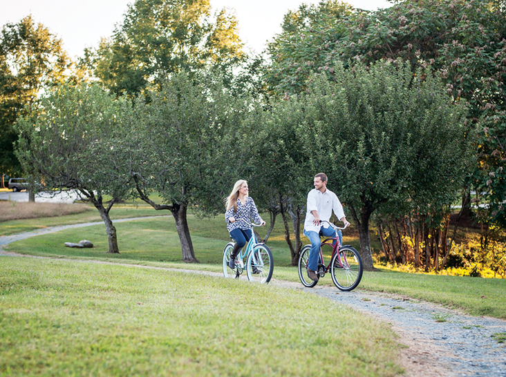A man and a woman ride bicycles on a winding path through a park with lush green grass and trees. The man wears a white shirt and the woman wears a patterned blouse. The sun is shining, creating a warm and serene atmosphere. Fearrington Village