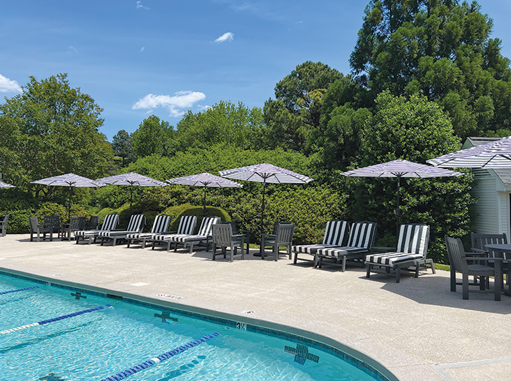 A poolside area featuring a row of black and white striped lounge chairs under matching umbrellas, set beside a swimming pool. The pool has swim lanes visible, and the surrounding area is lush with green trees and shrubs. The sky is clear and blue. Fearrington Village