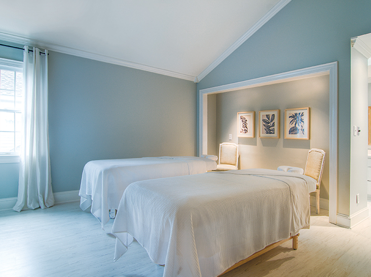 Bright, minimalist spa room with two massage tables covered in white linens. The room has soft blue walls, white curtains, and a high ceiling. Three botanical prints in white frames are displayed on the wall, and two chairs are placed near the tables. Natural light streams through a window. Fearrington Village