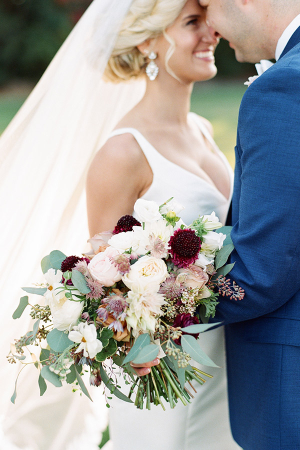 A bride and groom share an intimate moment on their wedding day. The bride, in a white dress and veil, holds a bouquet of various flowers including roses and dahlias. The groom is dressed in a blue suit. Both are smiling, and the background is blurred greenery. Fearrington Village