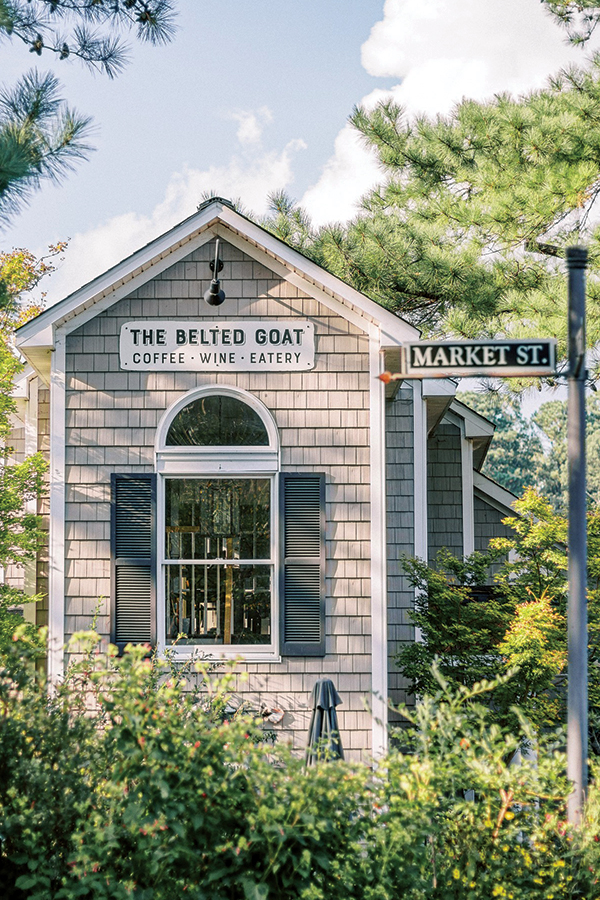 A charming building with grey shingles houses "The Belted Goat," a coffee, wine, and eatery establishment. Situated at the corner of Market Street, the building has a central window with black shutters, surrounded by greenery and trees under a blue sky. Fearrington Village