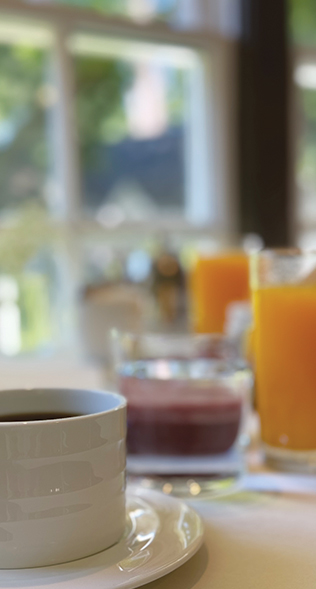 A close-up of a white coffee cup on a saucer at a breakfast table with beverages. In the background, there are glasses of orange juice and what appears to be a purple smoothie. The setting has a bright, blurred window view with greenery outside. Fearrington Village