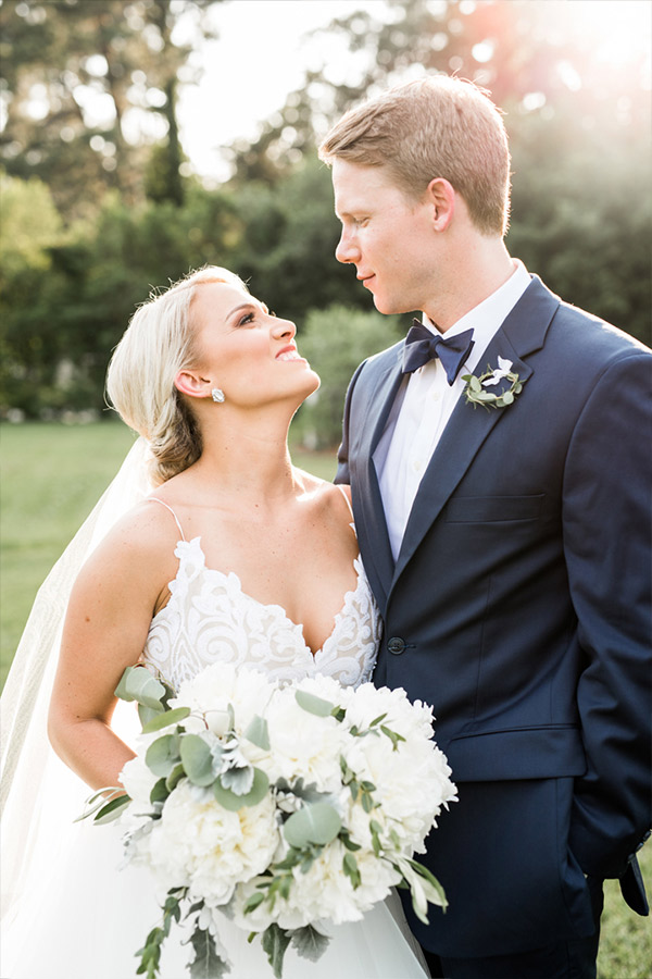 A newlywed couple gazes lovingly at each other in a sunlit outdoor setting. The bride, in a white gown with floral details, holds a bouquet of white flowers and greenery. The groom, in a navy blue suit and black bow tie, stands beside her with a serene expression. Fearrington Village