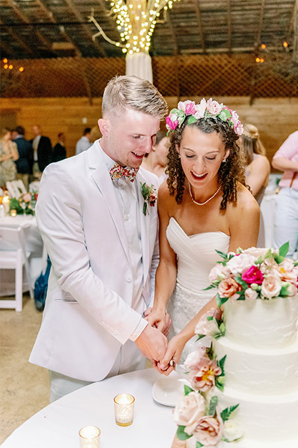 A bride and groom, dressed in a white wedding dress and white suit respectively, are cutting a white, multi-tiered wedding cake adorned with pink and white flowers. The bride wears a floral crown, and they are both smiling. Lights and guests are in the background. Fearrington Village