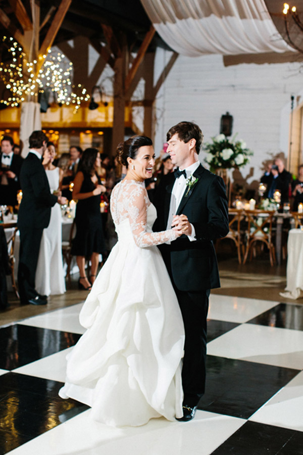 A bride and groom are dancing in a warmly lit reception hall. The bride is wearing a long-sleeved lace wedding gown, and the groom is in a black tuxedo. Other guests are seen in the background, some sitting and some standing, amidst decorations and string lights. Fearrington Village