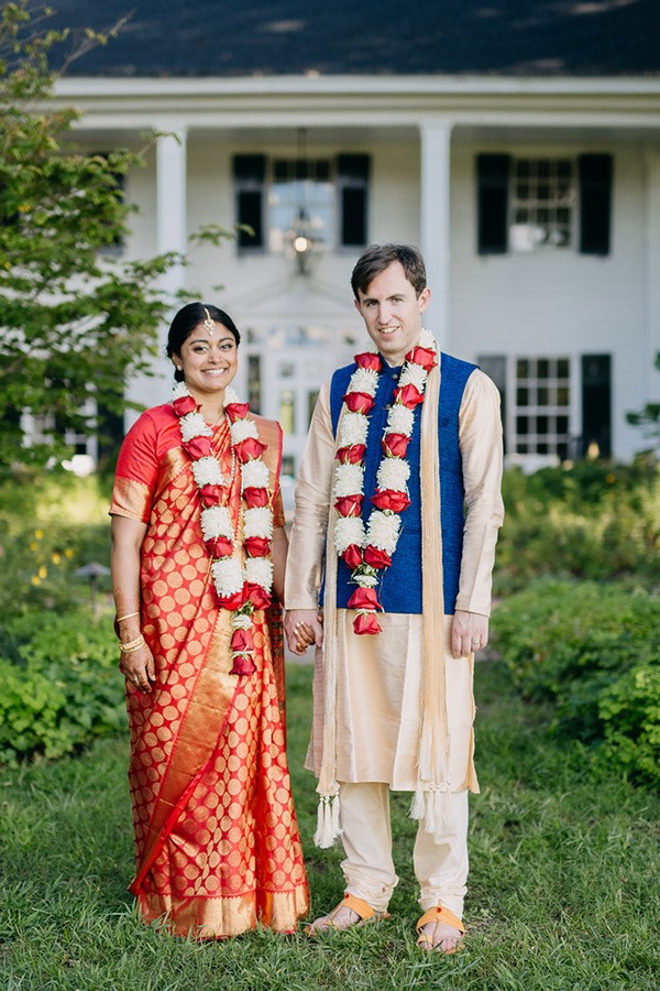 A couple stands outdoors in front of a building. The woman wears a red saree with gold patterns and a floral garland. The man wears a beige kurta with a blue vest and a matching garland. They hold hands and smile at the camera. Fearrington Village