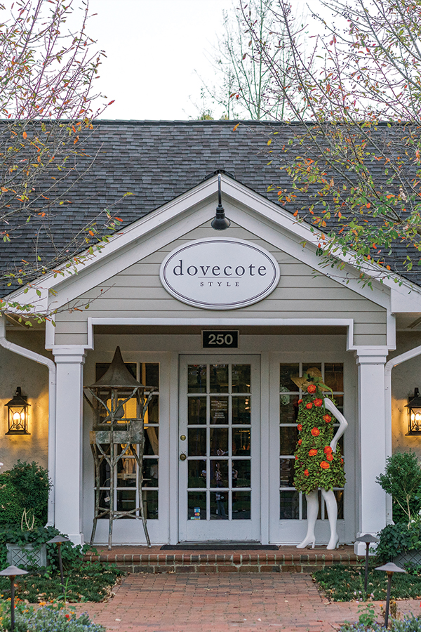 A quaint boutique named "dovecote style" with a shingled roof, located at 250. The entrance features a glass-paneled door, flanked by two decorative statues: one star-shaped and the other a mannequin adorned with greenery and red flowers. Fearrington Village