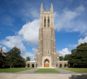 Duke Chapel. A tall, Gothic-style stone church tower stands prominently against a blue sky with scattered clouds, surrounded by trees and a manicured lawn. The central arched entrance is flanked by smaller arches and features wooden doors. Fearrington Village