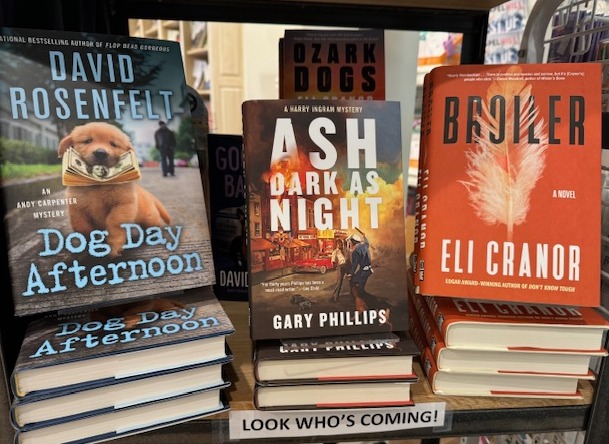 A bookstore display featuring stacks of three books: "Dog Day Afternoon" by David Rosenfelt with a dog on the cover, "Ash Dark As Night" by Gary Phillips with a dark cityscape, and "Broiler" by Eli Cranor with a fiery feather. A sign at the bottom reads "LOOK WHO’S COMING!. Fearrington Village