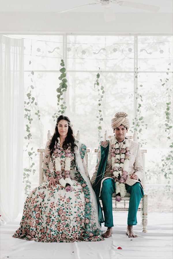 A bride and groom sit side by side on ornate chairs. Both wear traditional attire; the bride is in a floral dress with a green dupatta and floral garlands, the groom in a teal outfit with a turban and floral garlands. Green foliage decorates the background. Fearrington Village