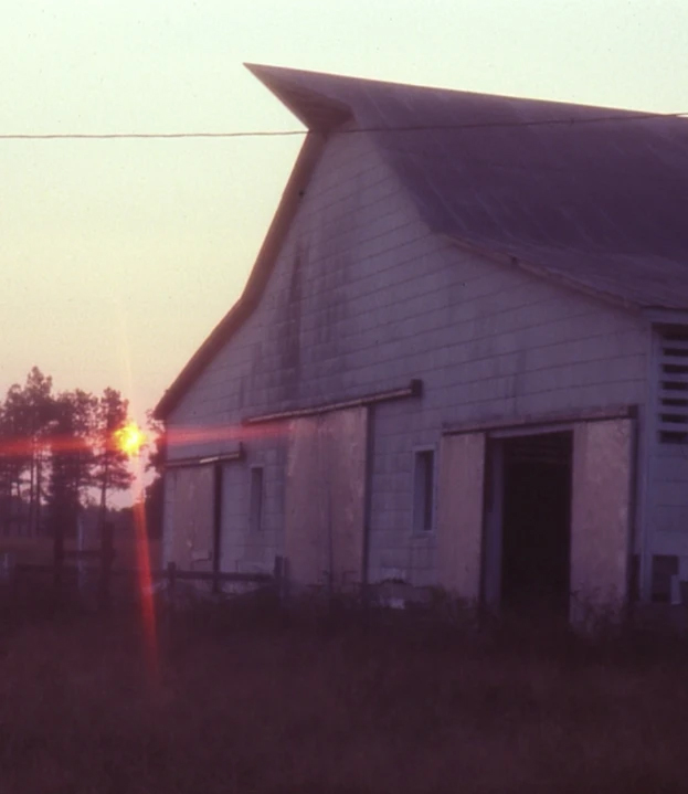 A sunset casts an orange glow through the silhouette of a rustic old barn with a distinctively pointed roof. The barn is in a rural landscape with trees in the background and an open doorway revealing its interior. The scene is serene and calm. Fearrington Village
