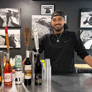 A person with long hair pulled back and wearing a cap and black shirt stands behind a bar counter. The countertop displays various bottles and cans of alcoholic beverages. Behind the person are black-and-white photos of animals on the wall. Fearrington Village