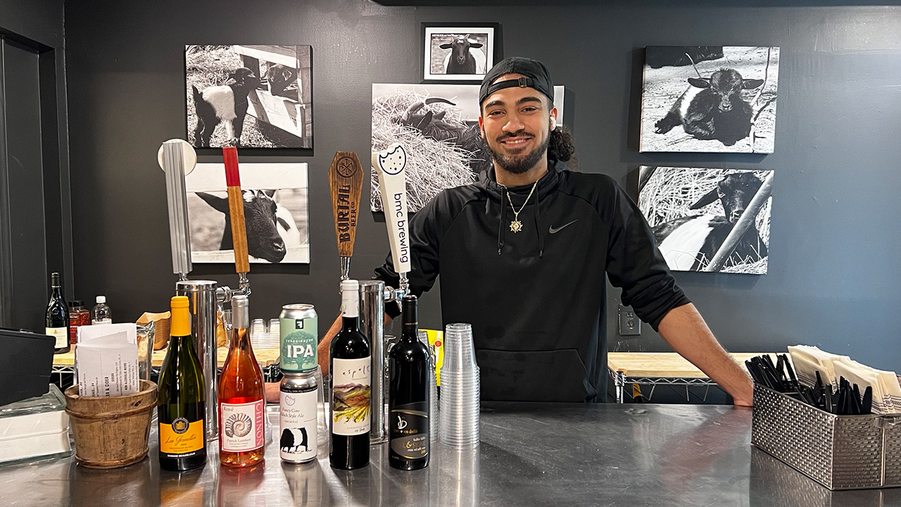 A person with long hair pulled back and wearing a cap and black shirt stands behind a bar counter. The countertop displays various bottles and cans of alcoholic beverages. Behind the person are black-and-white photos of animals on the wall. Fearrington Village