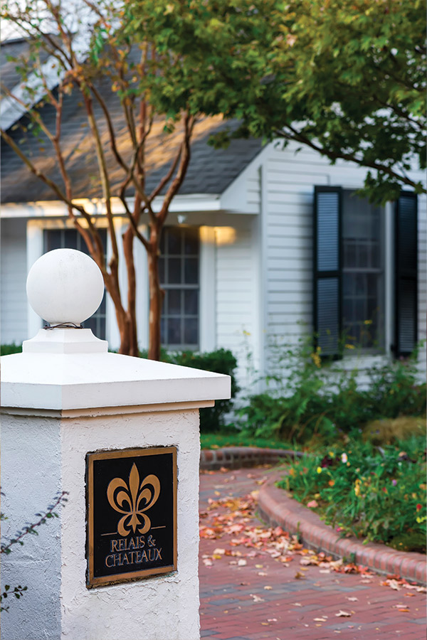 A white pillar with a plaque reading "Relais & Chateaux," featuring a golden fleur-de-lis symbol, stands in front of a charming white house with dark shutters and lush greenery. The path is lined with fallen autumn leaves. Fearrington Village