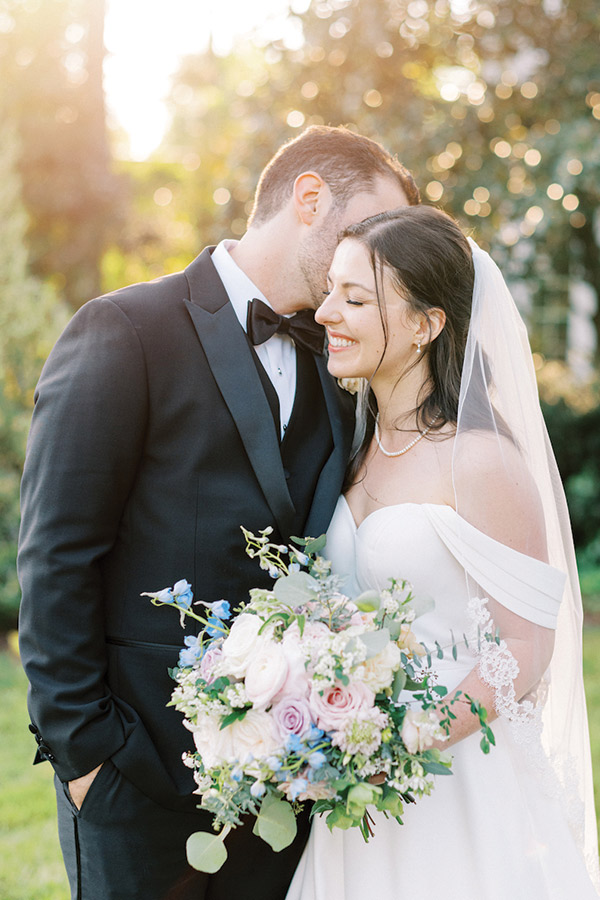 A bride in a white off-the-shoulder gown and veil smiles while holding a bouquet of flowers. She is standing close to a groom in a black suit and bow tie, who is leaning in to kiss her forehead. They are outdoors with greenery in the background. Fearrington Village