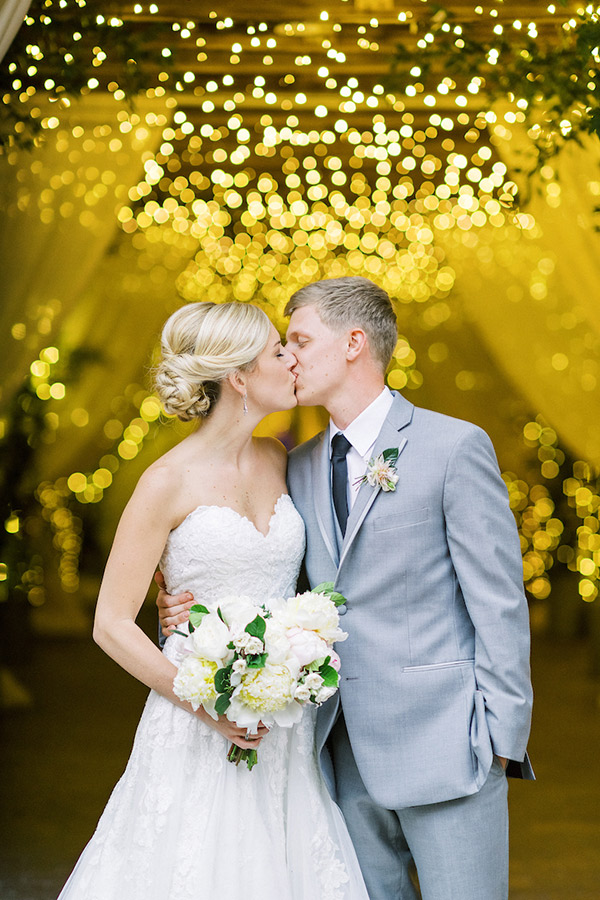 Bride and groom share a kiss under a canopy of twinkling lights. The bride, in a strapless white lace gown, holds a bouquet of white flowers. The groom, in a light gray suit with a black tie, gently embraces her. The background is filled with warm, golden fairy lights. Fearrington Village