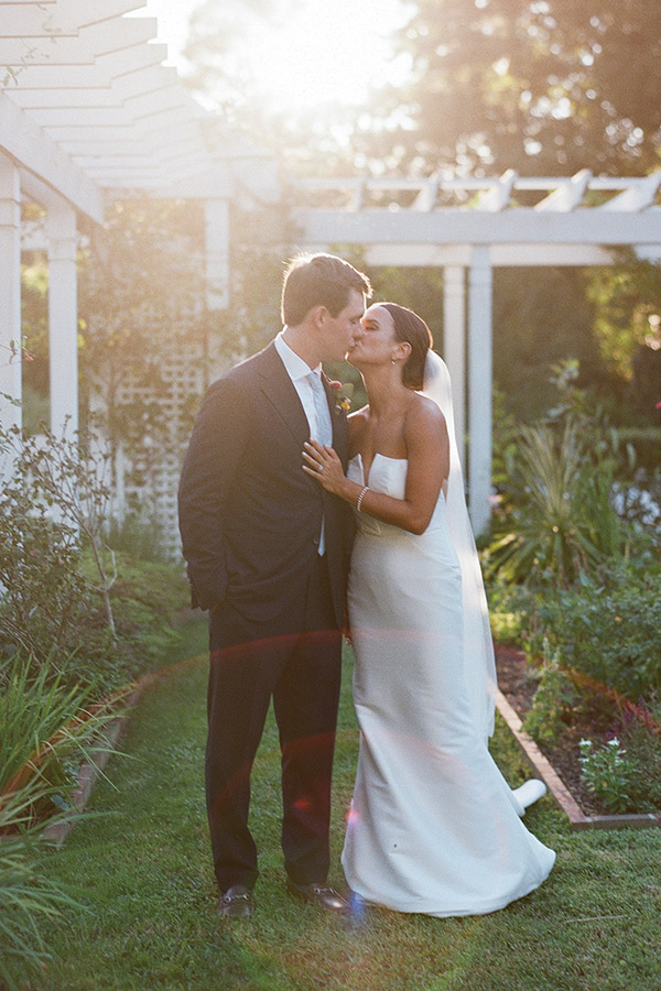 A bride and groom share a kiss in a sunlit garden. The groom wears a dark suit and a tie, the bride wears a strapless, white wedding gown with a veil. They stand under a white pergola surrounded by lush greenery, with sunlight creating a soft, romantic glow. Fearrington Village